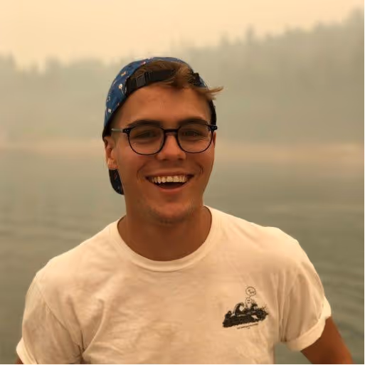 A young man with short, light brown hair is smiling widely, looking directly at the camera. He is wearing dark-rimmed glasses, a blue patterned baseball cap backward, and a white t-shirt with a dark graphic print on it. The background appears to be a misty or hazy outdoor scene with a body of water and faint trees, suggesting an overcast day or perhaps smoke/fog.