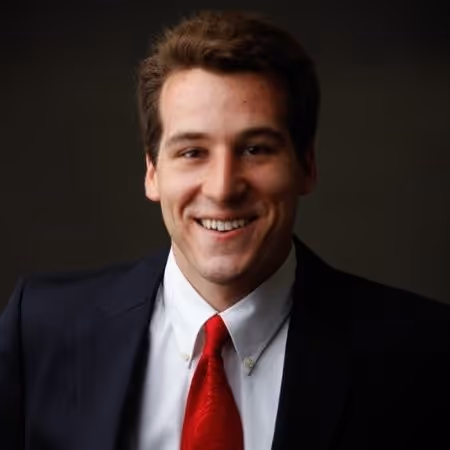 A professional headshot of a young man with short brown hair, smiling broadly at the camera. He is wearing a dark suit jacket, a white collared shirt, and a bright red tie. The background is a solid, dark gray or black.