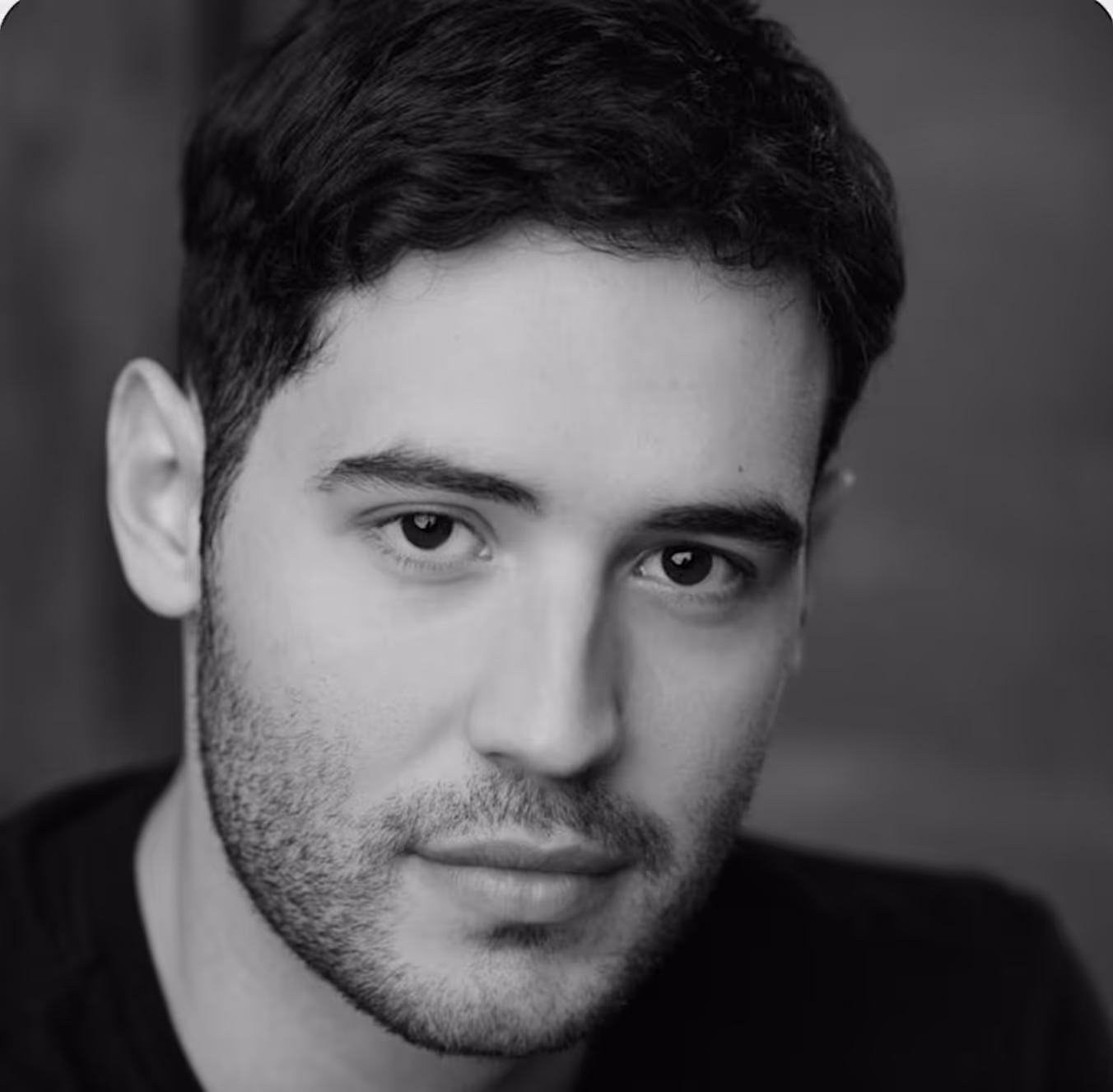 A black and white close-up headshot of a man with dark, short, curly hair and dark eyes. He has a beard and mustache, and is looking directly at the camera with a serious or neutral expression. The background is softly blurred.