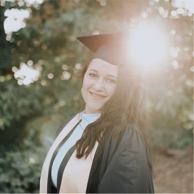 A young woman in graduation attire, including a black cap and gown with a light-colored stole, is smiling and looking at the camera. She has long, dark hair and hoop earrings. The sun is brightly flaring behind and to the right of her, creating a strong lens flare and backlighting. The background is blurred greenery.