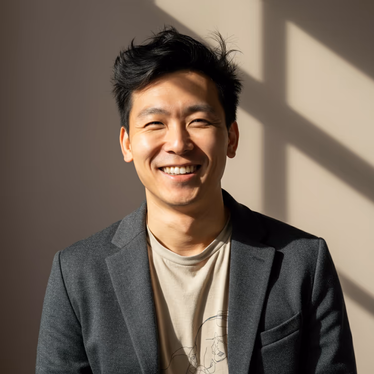 This is a bright and engaging headshot of a man in a modern, casual-professional style. He is wearing a charcoal gray blazer over a light beige t-shirt with a subtle graphic, with his dark hair styled in a slightly tousled manner. The portrait is taken against a simple, light-colored wall, using a strong, natural light source from the side which casts a dramatic shadow of a window frame diagonally across the background. This lighting technique highlights his genuine, broad smile and emphasizes a warm, confident, and approachable mood.
