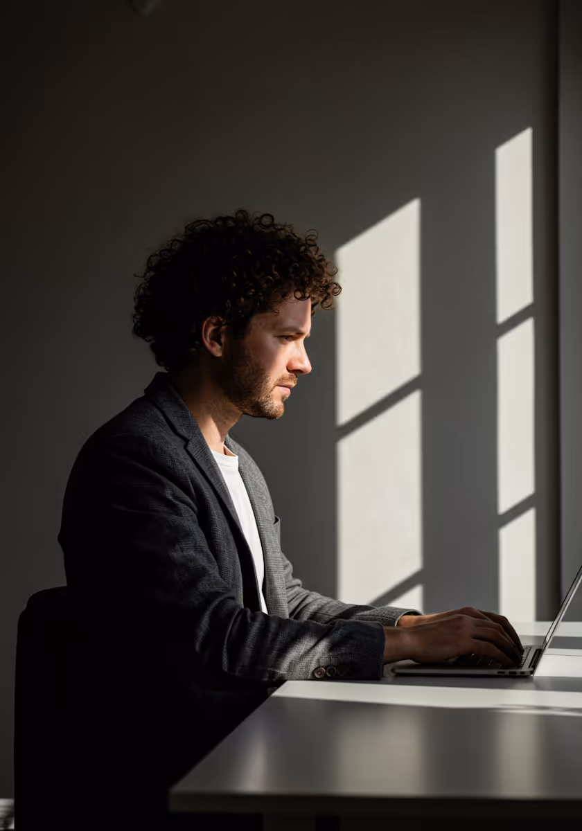 
This photograph features a man with curly hair and a beard sitting in profile, deeply focused on working on a laptop. He is dressed in a charcoal gray blazer over a white shirt. The image is characterized by dramatic, high-contrast lighting: a strong beam of natural light from a window strikes the right side of the frame and the man's face, creating a sharp geometric shadow on the plain, dark wall behind him and leaving much of his figure in deep shadow. This moody, cinematic lighting emphasizes his concentration and gives the scene an intense, contemplative atmosphere, suggesting he is engaged in private, focused work in an office or home setting.