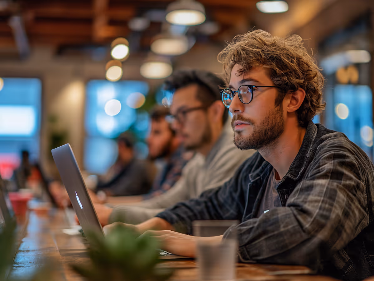 
This compelling photograph captures a row of focused individuals working on laptops in what appears to be a shared workspace, co-working facility, or modern café. The foreground is dominated by a young man with curly blonde hair, glasses, and a beard, dressed in a plaid shirt, intensely concentrating on his computer screen. The image uses a shallow depth of field, blurring the other people and the background to emphasize his focused work. The warm, moody lighting from the overhead fixtures and the exposed brick or industrial ceiling creates a cozy, yet productive and busy atmosphere characteristic of modern digital work environments.