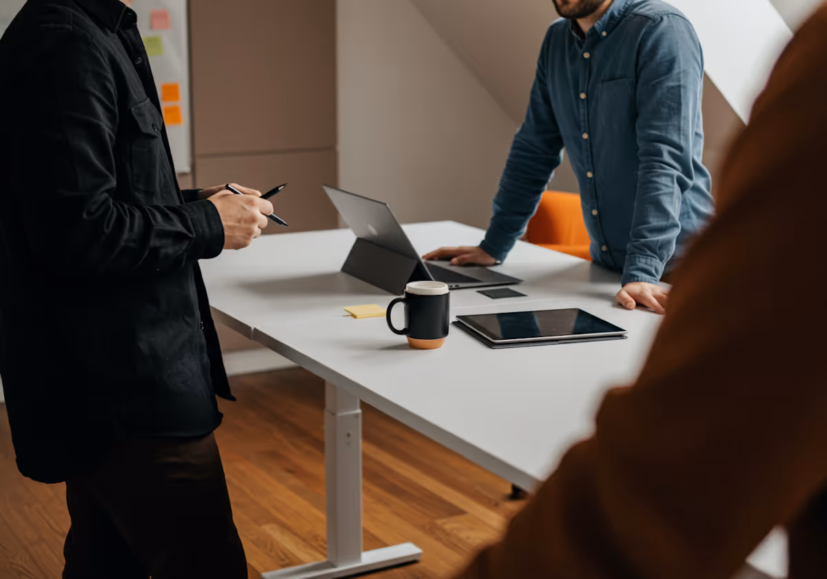 
This close-up image captures a moment of focused digital collaboration between colleagues around a clean, white adjustable desk. The scene shows at least three people, primarily focusing on the hands and upper torsos of two men. The man on the left, dressed in a black shirt, stands holding a pen, suggesting he is taking notes or preparing to annotate. The man on the right, wearing a blue denim-style shirt, leans on the desk near a laptop and a tablet, indicating an active discussion about the content displayed on the screens. A distinctive two-toned mug sits on the table between the devices. The setting, with its slanted attic ceiling and wooden floor, suggests a modern, casual, yet highly productive office or startup environment.