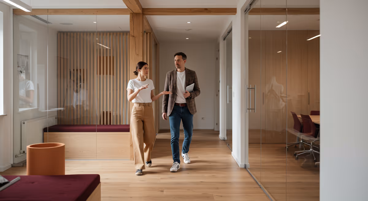 
This full-shot image captures two colleagues walking and discussing a topic as they move through a bright, modern, and beautifully designed office corridor. The woman on the left, wearing a white t-shirt and light brown trousers, is actively engaged in conversation, using an expressive hand gesture to emphasize her point. The man, dressed in a brown tweed blazer, jeans, and white sneakers, walks beside her, holding a tablet or notebook. The architecture is characterized by extensive use of wood paneling and beams contrasted with clear glass walls and doors, which define the meeting rooms and private offices, creating an open yet structured workplace with a warm, contemporary aesthetic.