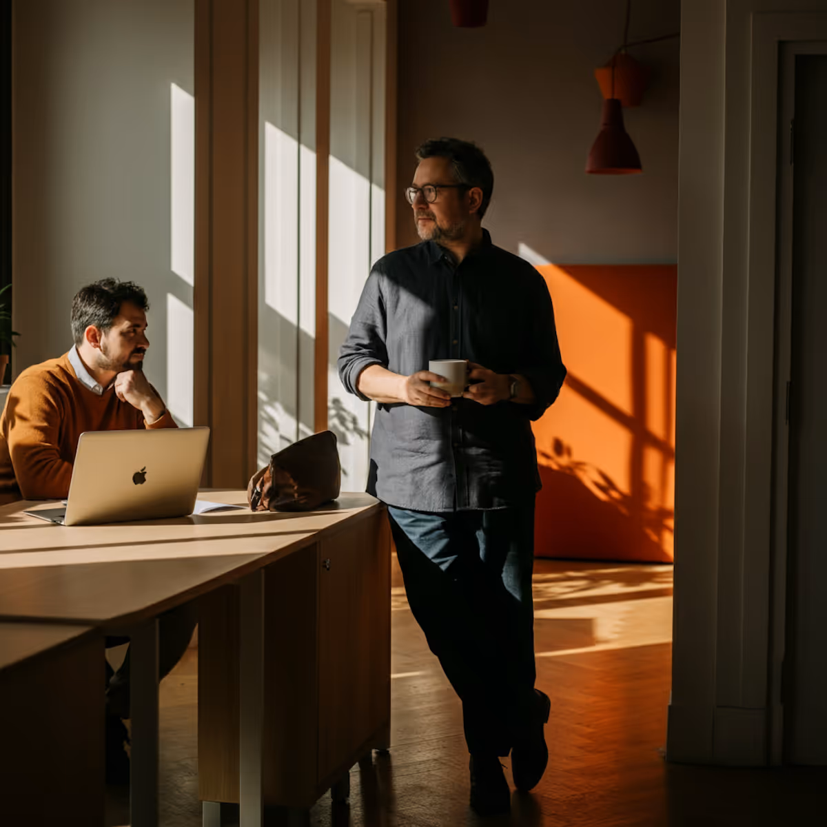 
This striking image captures two men in a moment of thought or casual interaction within a stylish office bathed in dramatic, warm, natural light. In the foreground, one man, wearing a dark button-up shirt and glasses, stands leaning against a wooden desk, holding a coffee mug and looking reflectively off-camera toward the window light. In the background, a second man in an orange sweater sits at the desk, looking at the first man while a MacBook and a leather bag rest near him. The room features a contrasting color palette, with the bright orange wall in the distance and the geometric patterns of sunlight and shadow cast across the floor and paneling, suggesting a break or a pause in a creative work session.
