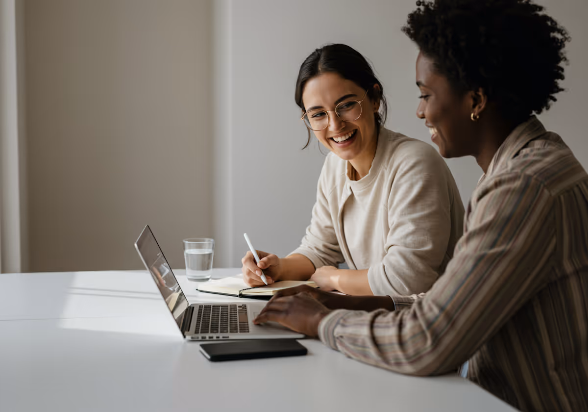 
This engaging image captures two women collaborating with visible enthusiasm in a brightly lit, minimalist office setting. The woman on the left, wearing glasses and a cream-colored sweater, is smiling genuinely while taking notes in a notebook with a pen. The woman on the right, with curly hair and a striped, earth-toned blouse, is actively working on a laptop, seemingly sharing content with her colleague. The clean, white desk and neutral gray wall create a distraction-free background, emphasizing the positive and productive interaction between the two, which appears to be a highly successful and enjoyable moment of teamwork.