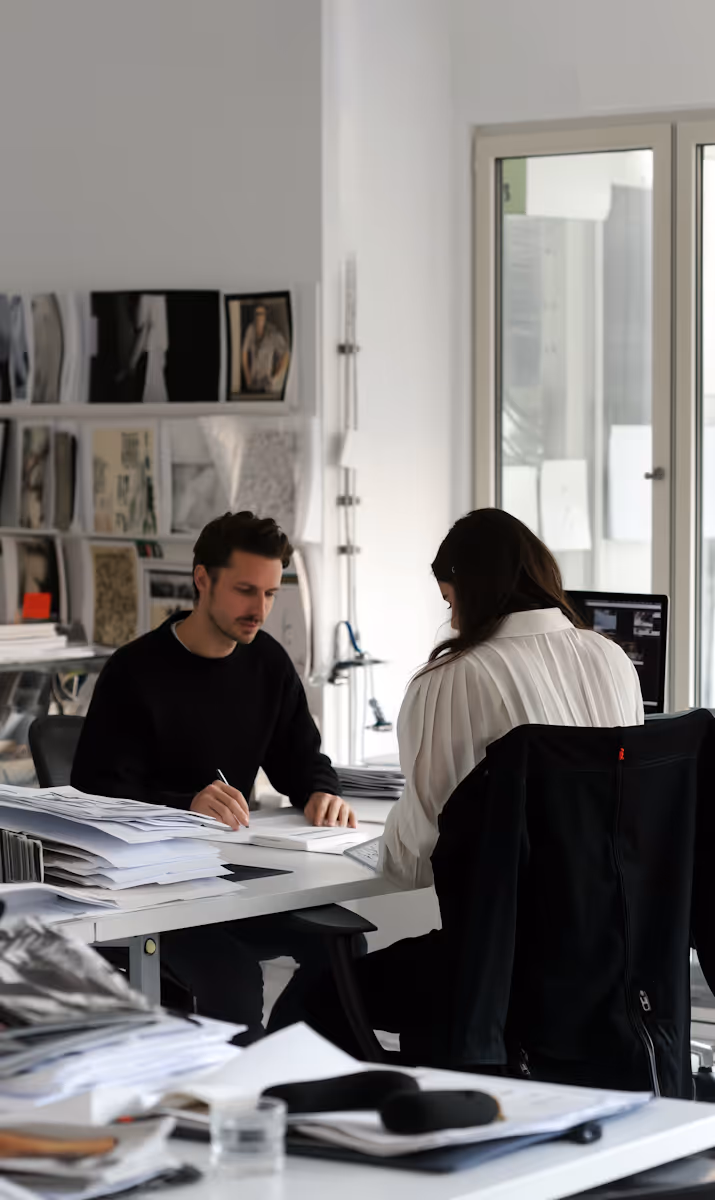 
This image portrays a focused moment of one-on-one collaboration within a bright, contemporary design studio or office. A man, wearing a black sweater, sits on the left intently writing or sketching on paper, surrounded by stacks of documents and papers. Opposite him sits a woman, her back mostly to the camera, distinguished by a white, pleated blouse, working at a computer screen. The environment is clearly a creative workspace, with shelves full of mood boards, sketches, and reference materials visible on the back wall, suggesting they are colleagues engaged in a detailed project review or design session.