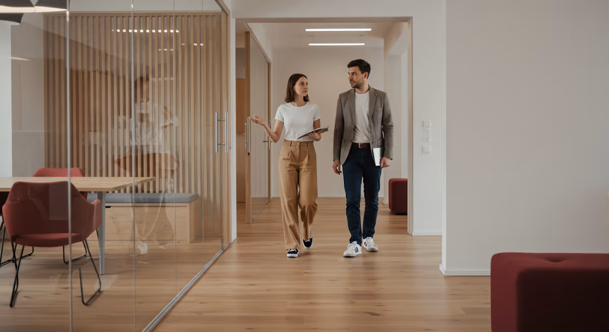 
This full-shot image captures two colleagues walking and discussing a topic as they move through a bright, modern, and beautifully designed office corridor. The woman on the left, wearing a white t-shirt and light brown trousers, is actively engaged in conversation, using an expressive hand gesture to emphasize her point. The man, dressed in a brown tweed blazer, jeans, and white sneakers, walks beside her, holding a tablet or notebook. The architecture is characterized by extensive use of wood paneling and beams contrasted with clear glass walls and doors, which define the meeting rooms and private offices, creating an open yet structured workplace with a warm, contemporary aesthetic.