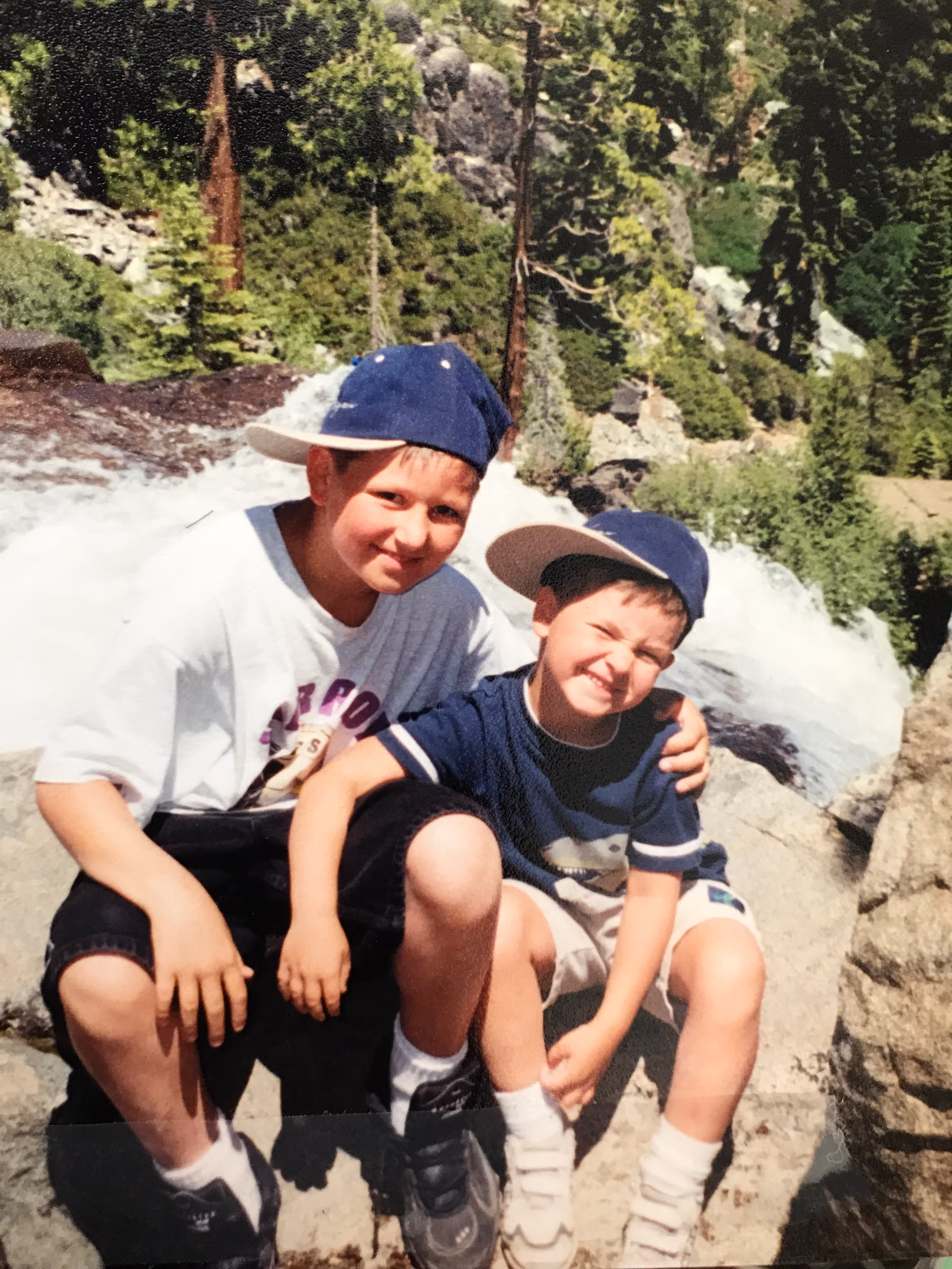 Two young boys, both wearing baseball caps, are sitting on rocks outdoors. The boy on the left, slightly older, is wearing a white t-shirt and dark shorts, with a blue cap on backward. He has his arm around the younger boy, who is wearing a dark blue t-shirt, light-colored shorts, and a blue cap also on backward. The younger boy is smiling broadly with squinted eyes. Behind them, there's a rocky, natural landscape with trees and what appears to be a stream or waterfall in the background. The photo has a slightly faded, vintage quality.