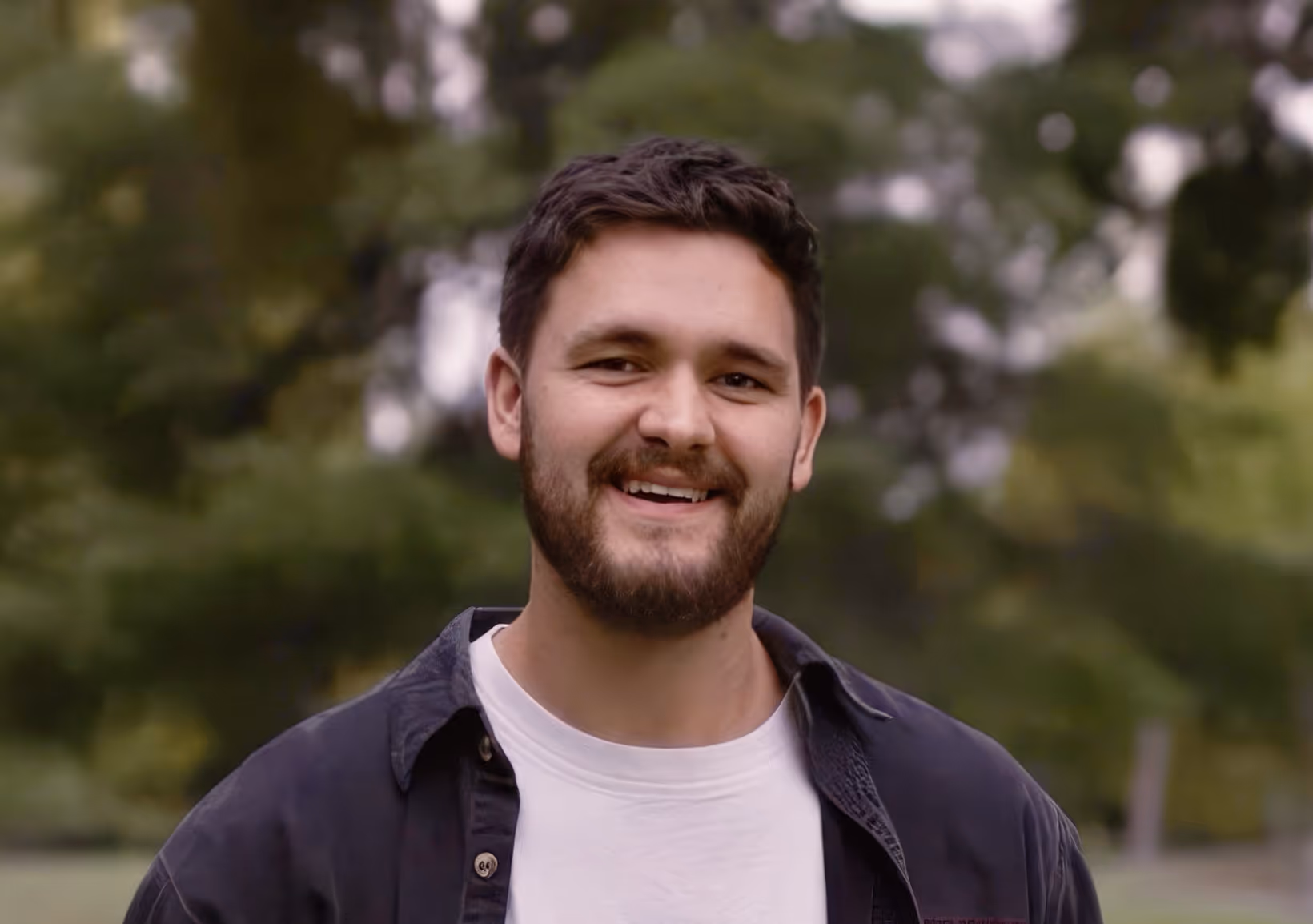 A headshot of a smiling man with dark hair and a beard, standing outdoors. He is wearing a white t-shirt underneath a dark, unbuttoned collared shirt or light jacket. The background is a softly blurred natural setting of dark green trees and foliage, giving the photo a candid and warm feeling.