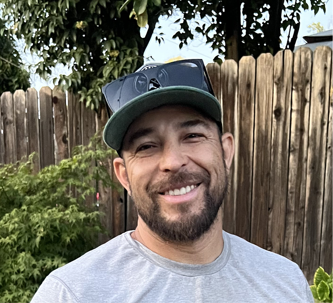 A smiling man with a beard and a baseball cap is shown in a headshot outdoors. A wooden fence and green foliage are blurred in the background.