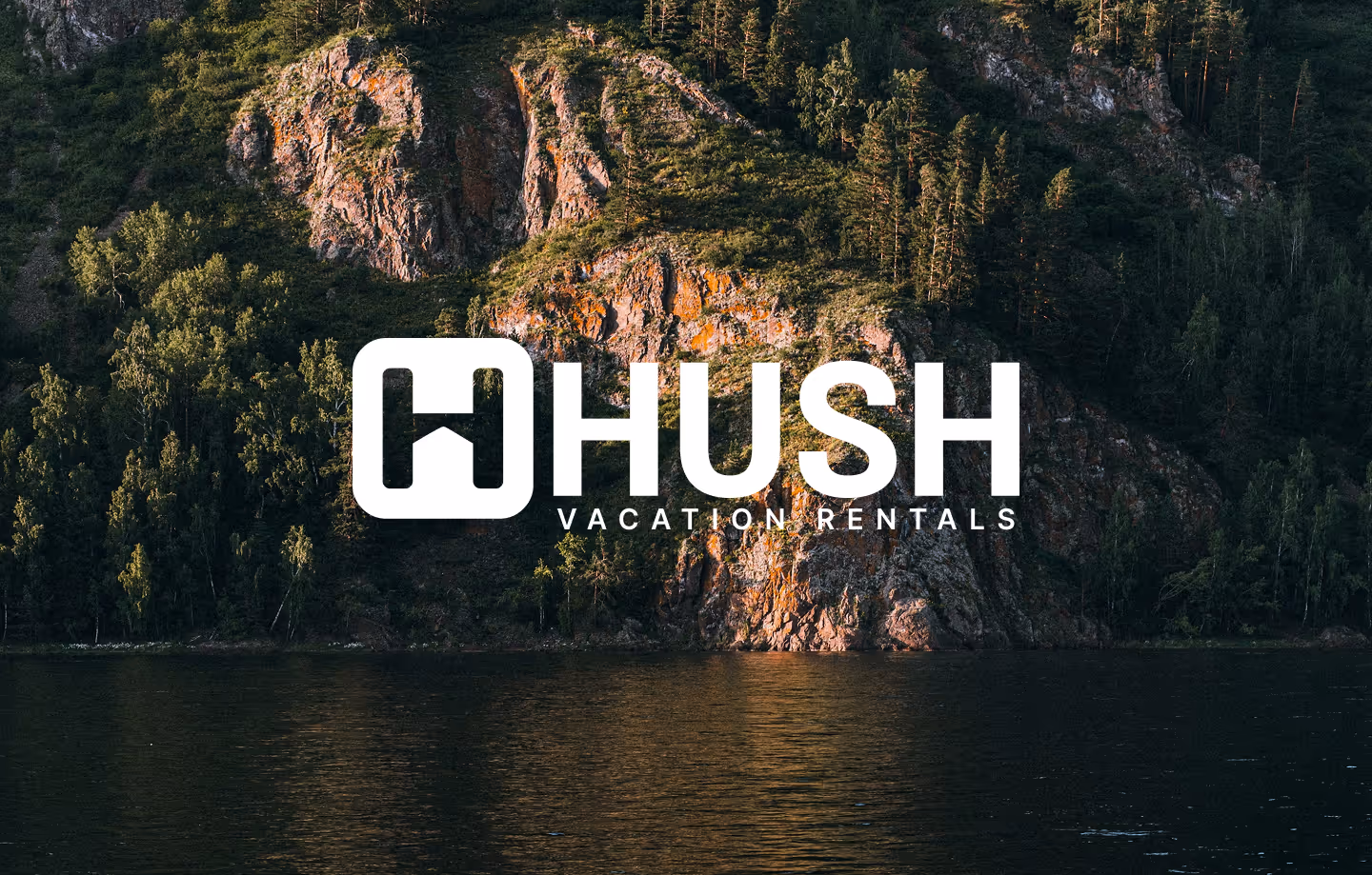 The "HHUSH VACATION RENTALS" logo in white is superimposed over a scenic landscape. The background features a large rocky mountain face covered with green trees, and a dark body of water in the foreground.