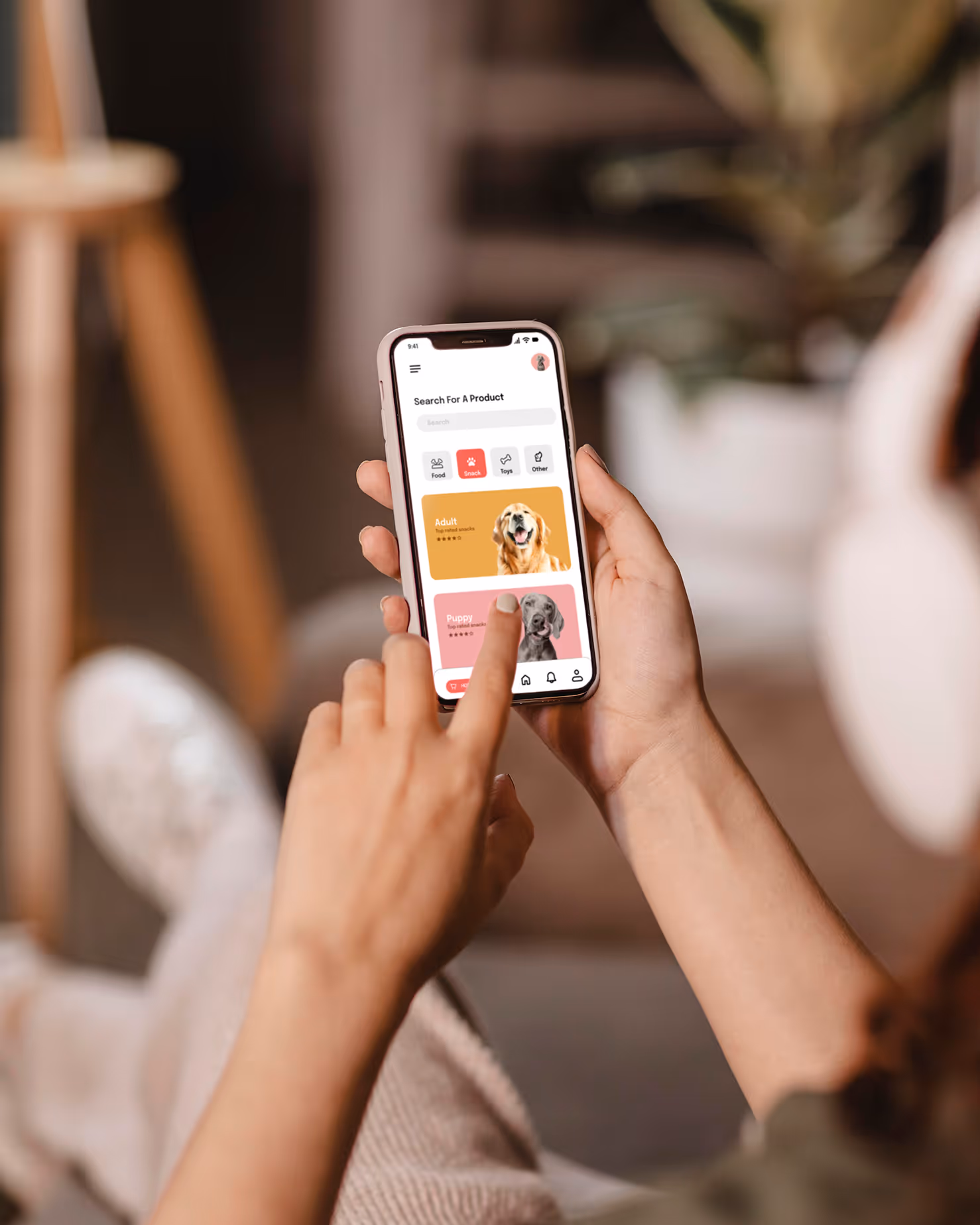 A person is holding a smartphone with both hands, using their thumbs to interact with the screen. The phone displays a "Search for a Product" interface with images of dogs and product categories like "Adult" and "Puppy," suggesting a pet-related app.