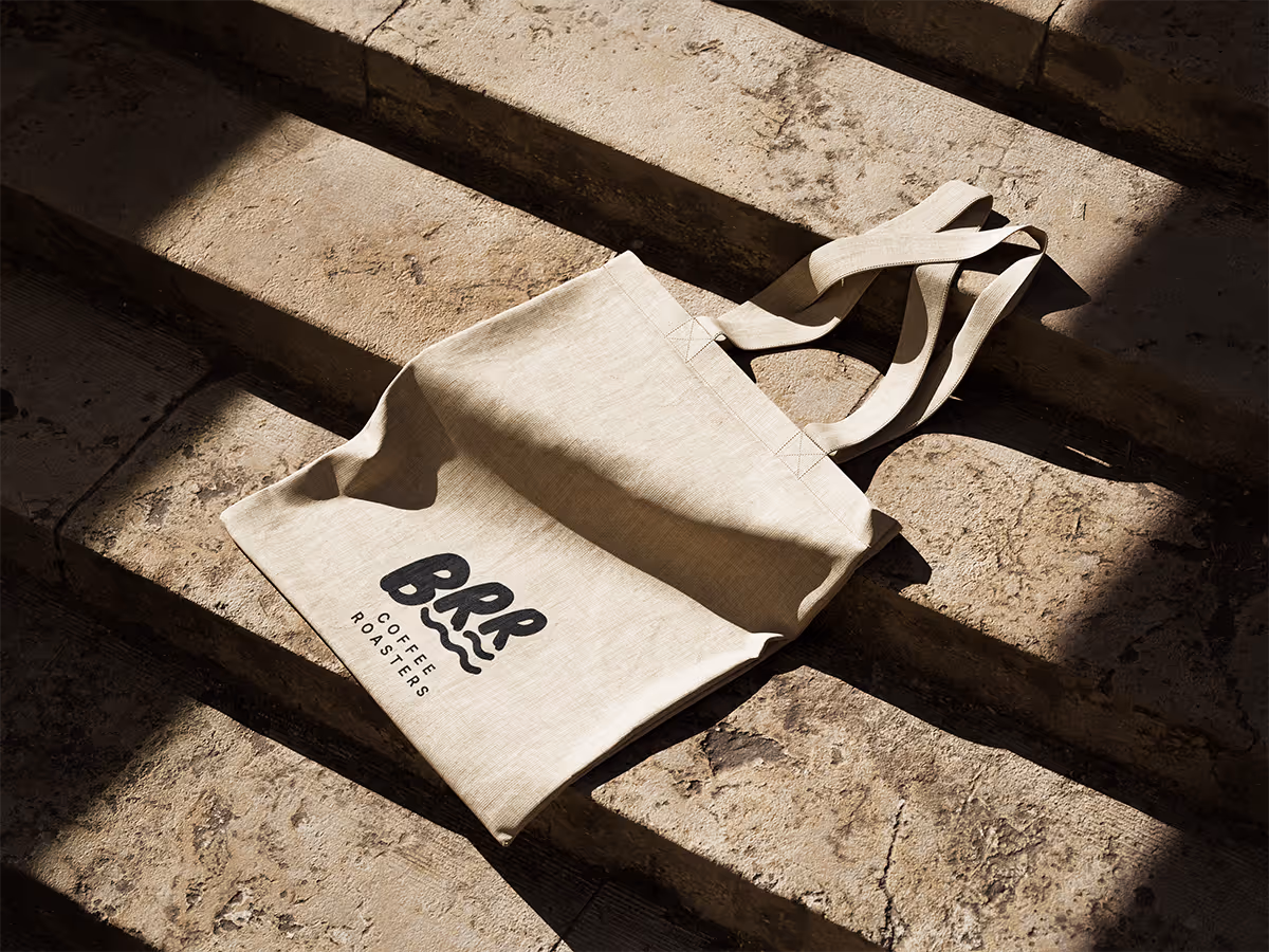 A light-colored canvas tote bag with a dark "BRR COFFEE ROASTERS" logo is lying on stone steps. Strong sunlight casts distinct shadows, highlighting the texture of the bag and steps.