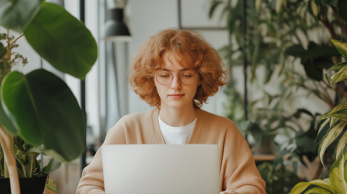 An image shows a person with curly reddish-brown hair and glasses, wearing a light brown cardigan, looking down at a laptop. They are surrounded by green plants.
