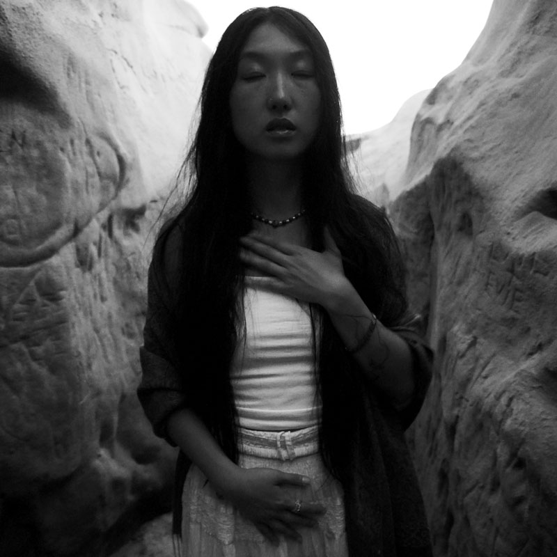 A smiling Korean woman leaning against a rock face on a beach.