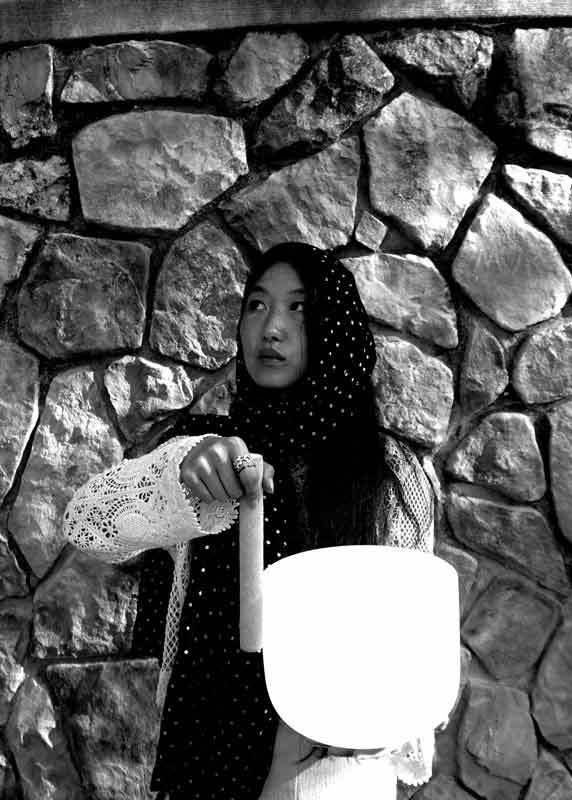 An Korean woman holding a singing bowl in front of a cobblestone wall.