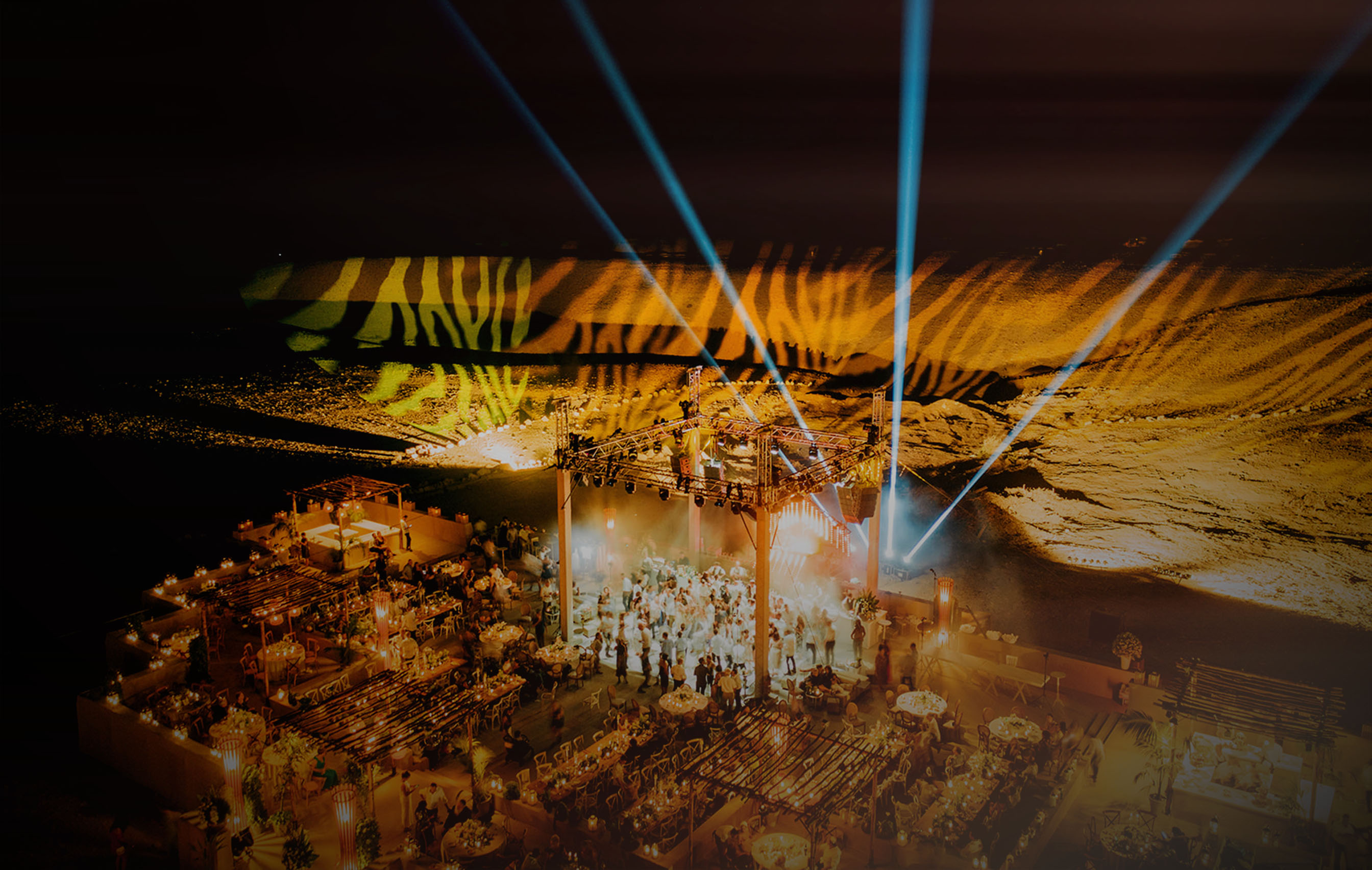 Elegant desert wedding in Israel, held at night with dramatic lighting projections on the sand, beams of light in the sky, and a vibrant celebration with dining tables, a dance floor, and guests enjoying the event.