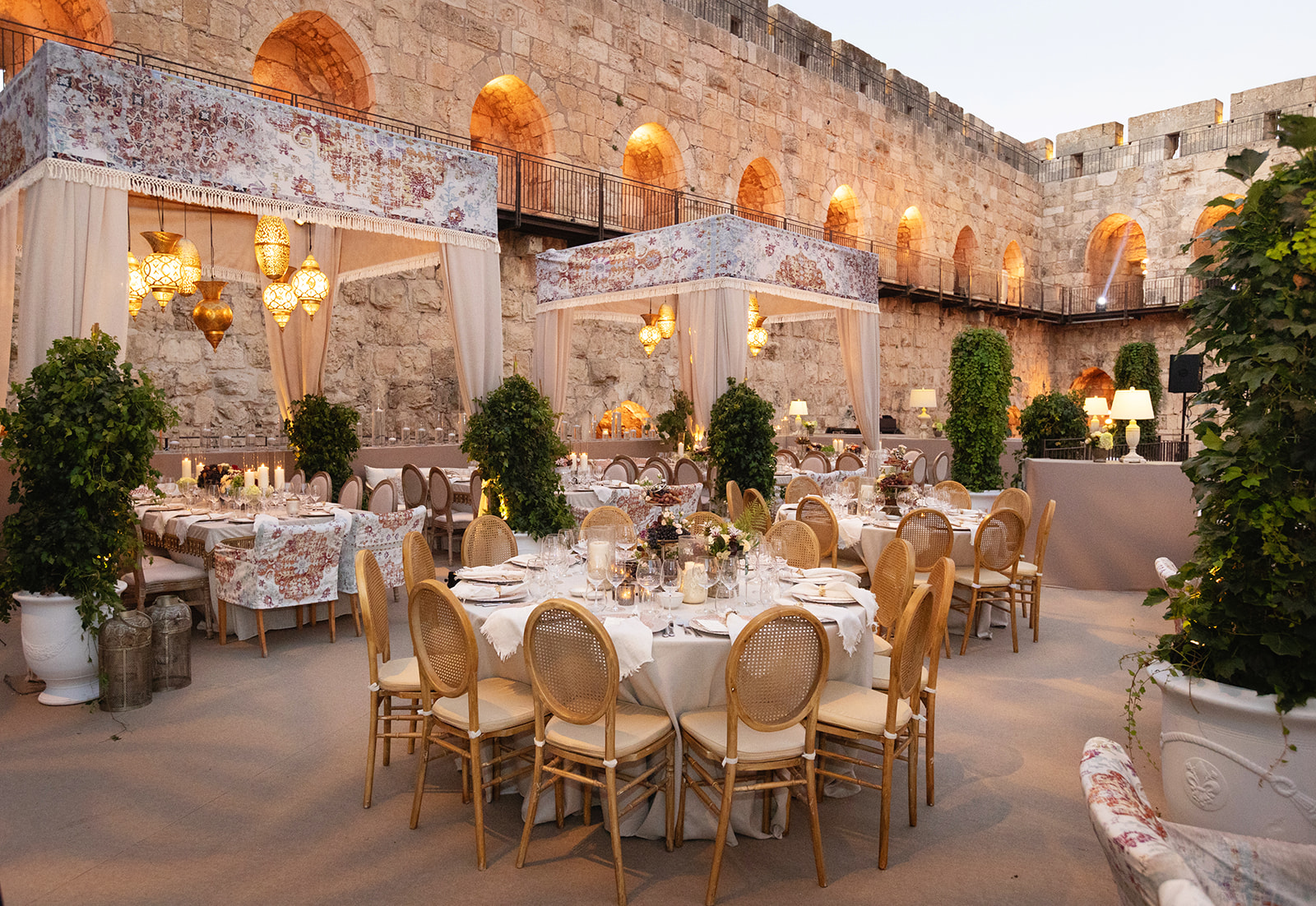 Elegant wedding setup inside the historic Tower of David in Jerusalem, featuring round dining tables, soft beige and floral fabrics, ambient lantern lighting, and the ancient stone walls glowing warmly in the evening light.