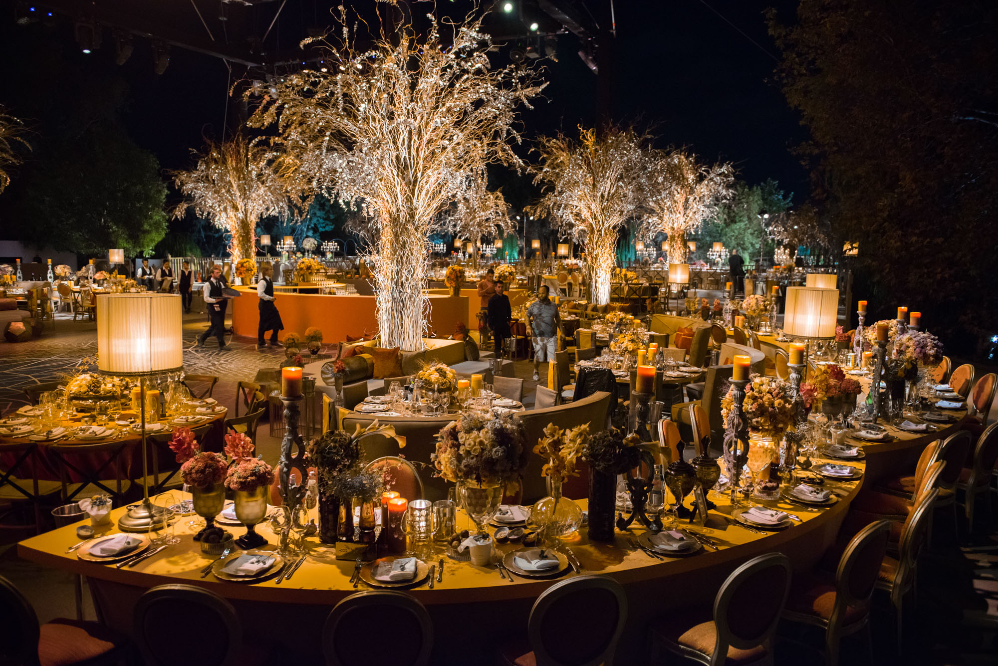 Lavish nighttime wedding reception at Ronit Farm in Israel, featuring dramatic illuminated tree installations, curved banquet tables with ornate place settings, warm candlelight, and lush floral arrangements under an open sky.