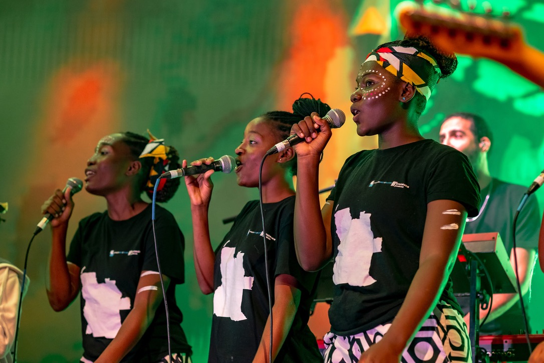 Three female vocalists perform passionately on stage, dressed in black t-shirts with white Africa-shaped prints and colorful head wraps, illuminated by vibrant green and orange stage lighting.
