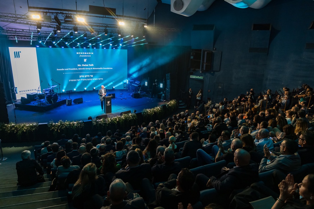 A large audience attentively watches a keynote speech at a Menomadin Foundation event, featuring a speaker on stage in front of a bright blue screen with the foundation’s branding, under professional stage lighting.
