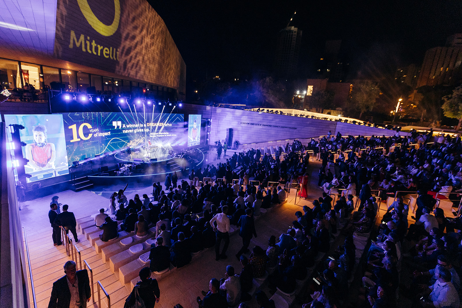 Wide-angle night view of an outdoor corporate event at the Jerusalem Theater, featuring a large crowd seated around a circular stage with dynamic lighting and digital screens displaying “10 Years of Impact” for Mitrelli.