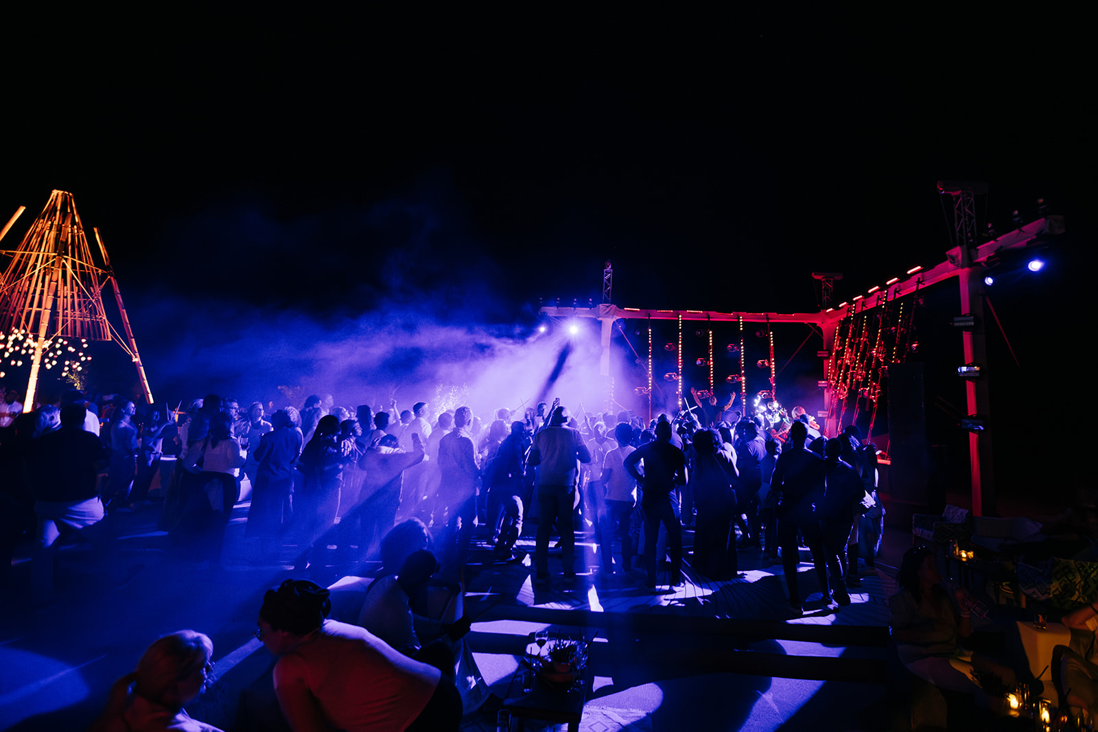 A wide shot of an outdoor night party bathed in blue and purple lights, with a crowd of people dancing and a stage setup in the background.