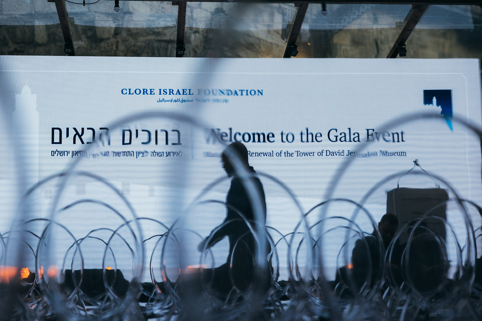 Silhouettes of crew members setting up in front of a large screen that reads “Welcome to the Gala Event” at the Tower of David Museum, seen through rows of transparent chairs.