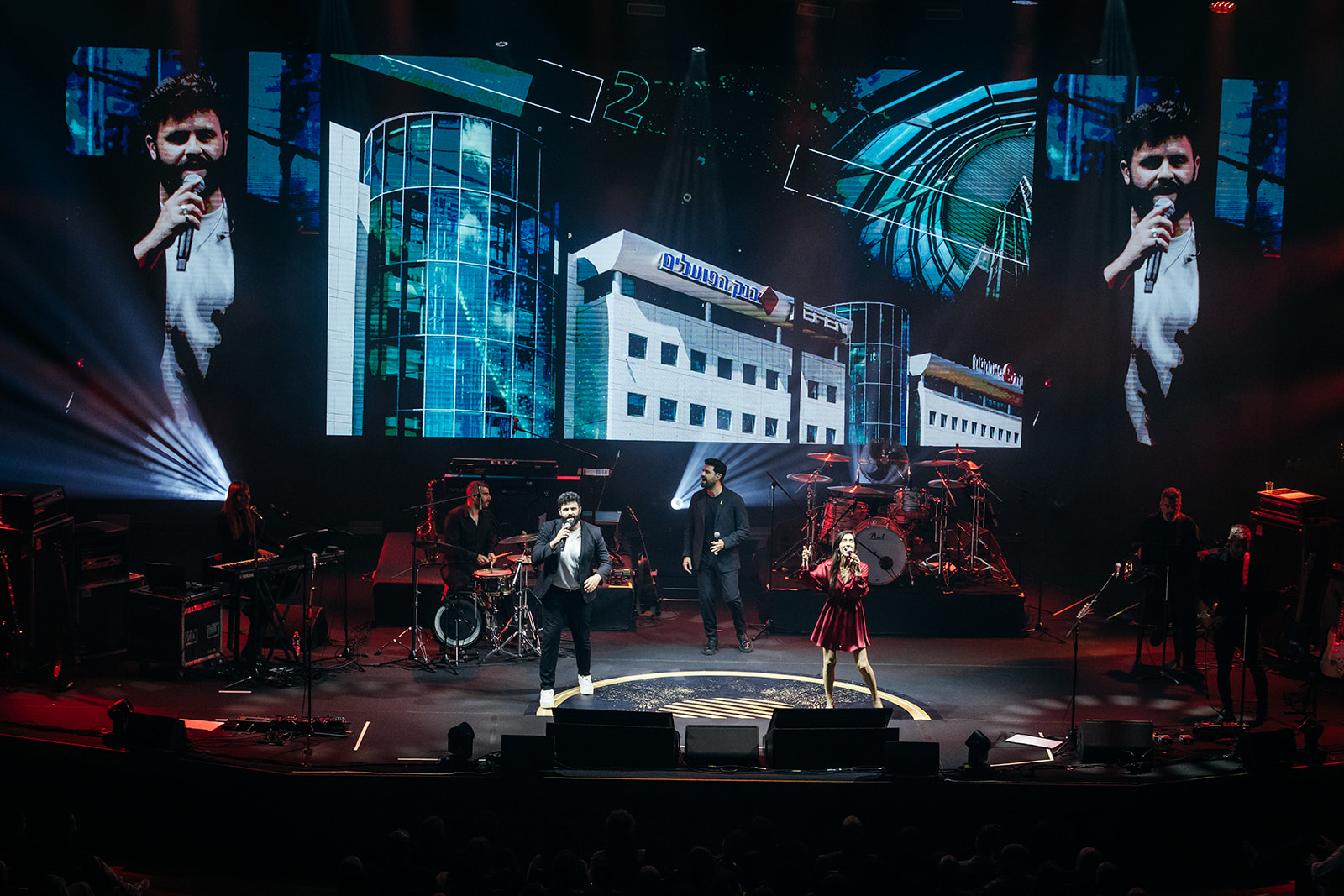 Wide stage view with musicians and large LED screens showing animated visuals of Amot buildings.