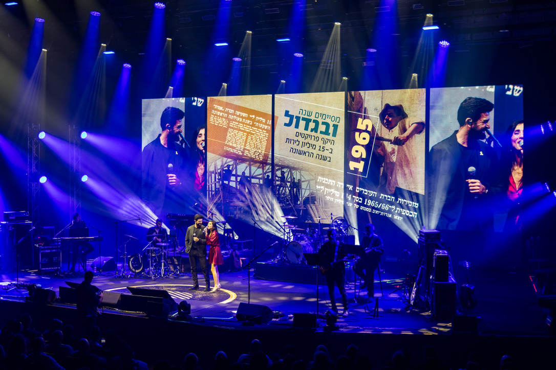 Live performance on stage with dramatic lighting and large LED screens displaying Amot's history and branding.