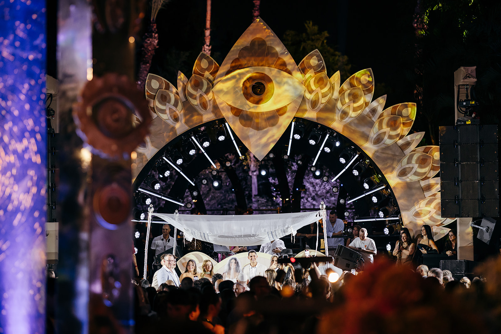 Couple under a white chuppah at their Israeli wedding, framed by a decorative eye-shaped stage structure and seated guests. Unique wedding venue Israel.