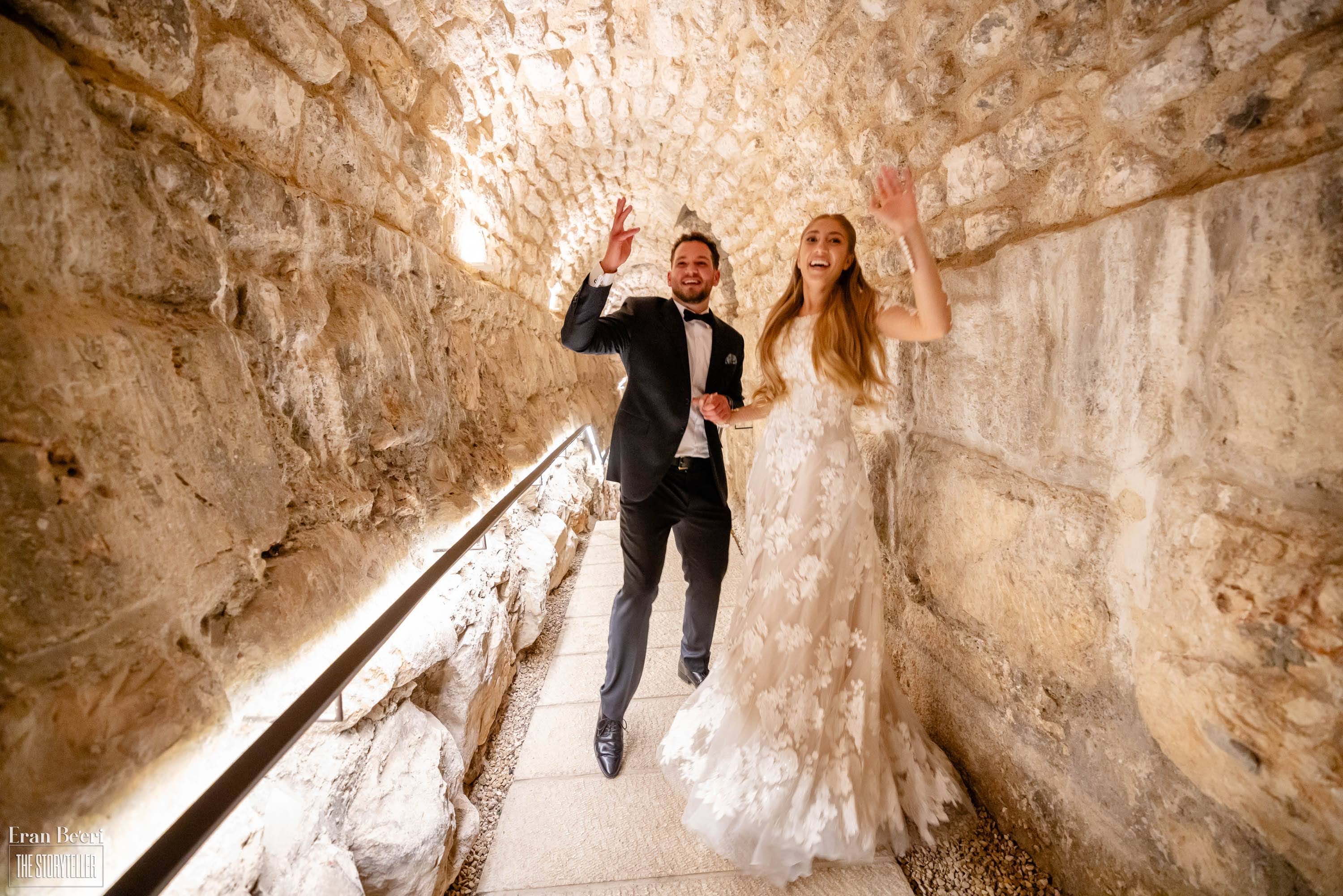 Bride and groom walking hand in hand through a warmly lit ancient stone passageway at the tower of david, smiling and waving toward the camera on their wedding day.