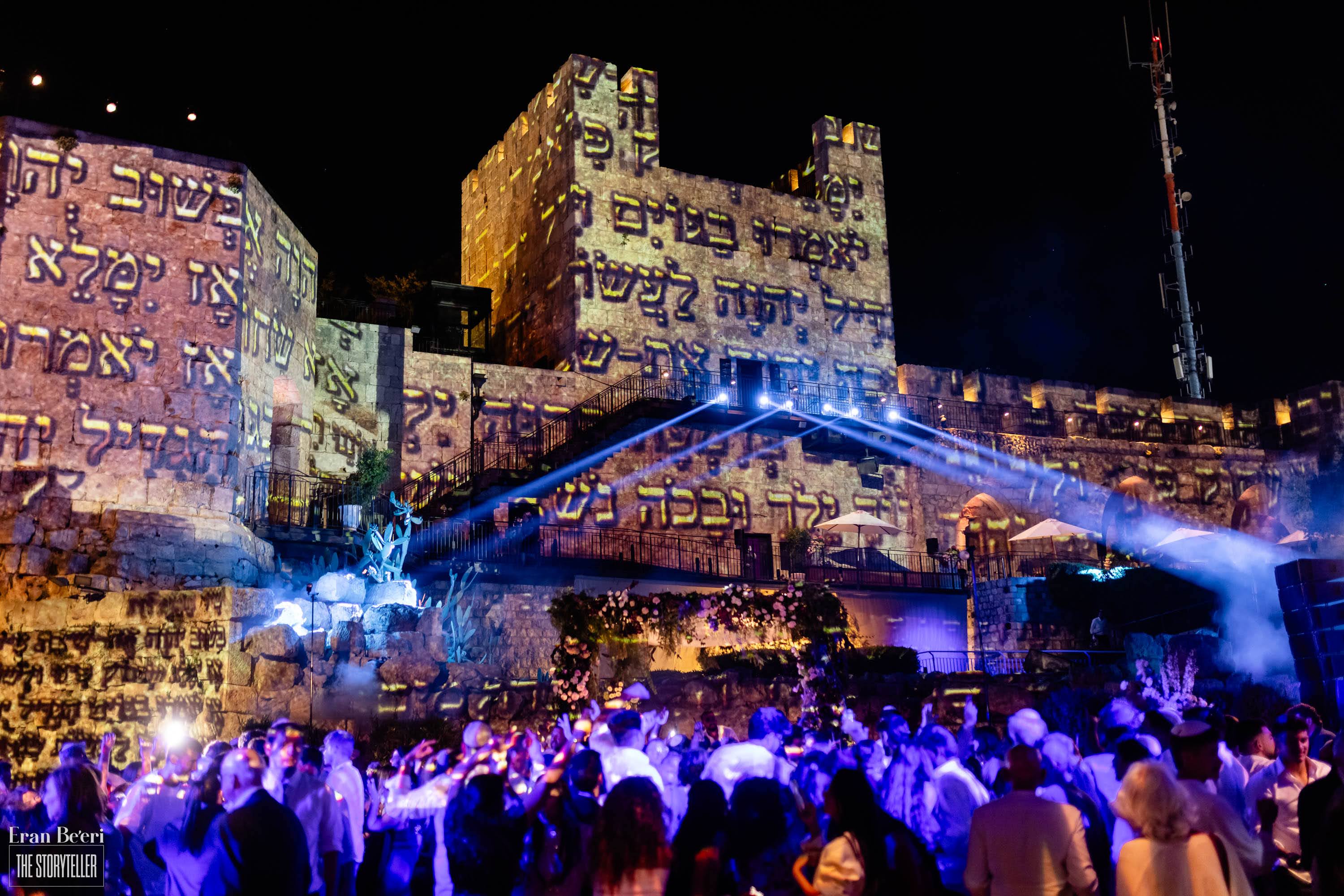 Nighttime wedding celebration with dramatic Hebrew text projections and blue lighting across the historic Tower of David walls in Jerusalem.