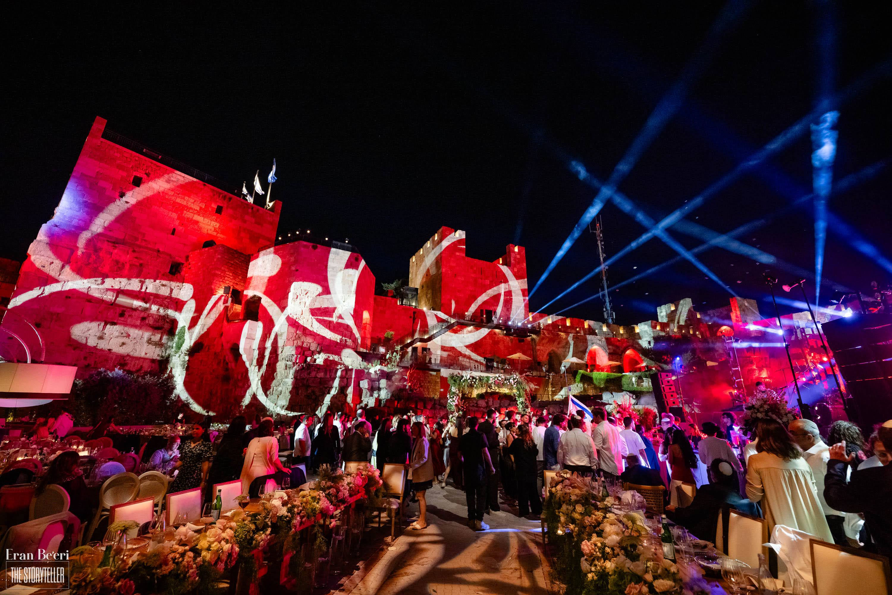 Spectacular nighttime wedding celebration with dramatic red projections and blue stage lighting across the historic Tower of David in Jerusalem.