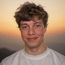 A young man with curly brown hair is smiling softly at the camera. He is wearing a white T-shirt and standing in front of a warm, hazy sunset with soft mountains in the background.