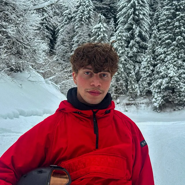 A young man in a red ski jacket stands in a snowy forest, holding a helmet and looking at the camera.