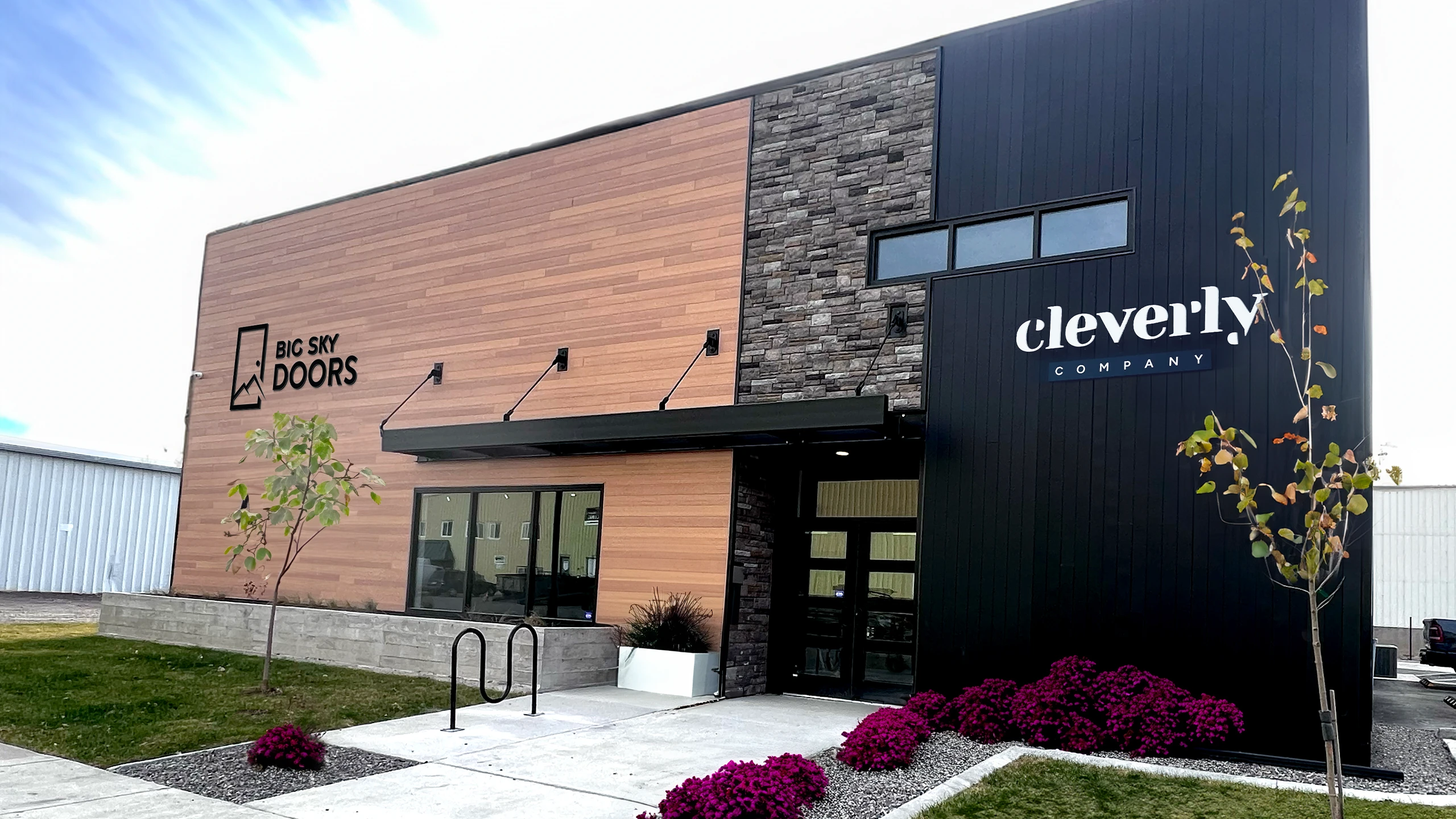 Exterior of The Cleverly Company headquarters, a modern commercial building featuring wood paneling, stone accents, and black vertical siding. The company’s bold white logo is displayed on the right side of the facade. The landscaped front entrance includes young trees, vibrant purple flowers, and a concrete walkway.