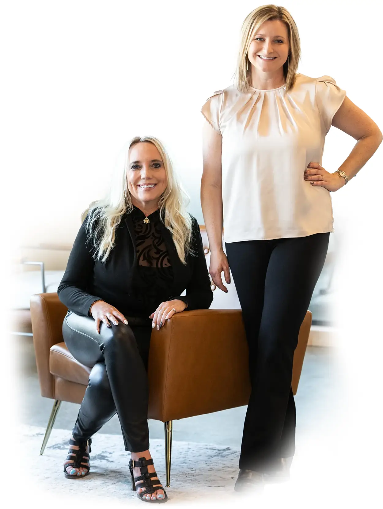 Portrait of Kally Amundsen and Jenn Fancher, co-founders of The Cleverly Company. Kally is seated in a modern tan leather chair, wearing a black top and leather pants, while Jenn stands beside her in a soft beige blouse and black pants. Both women are smiling confidently in a bright, modern office setting.