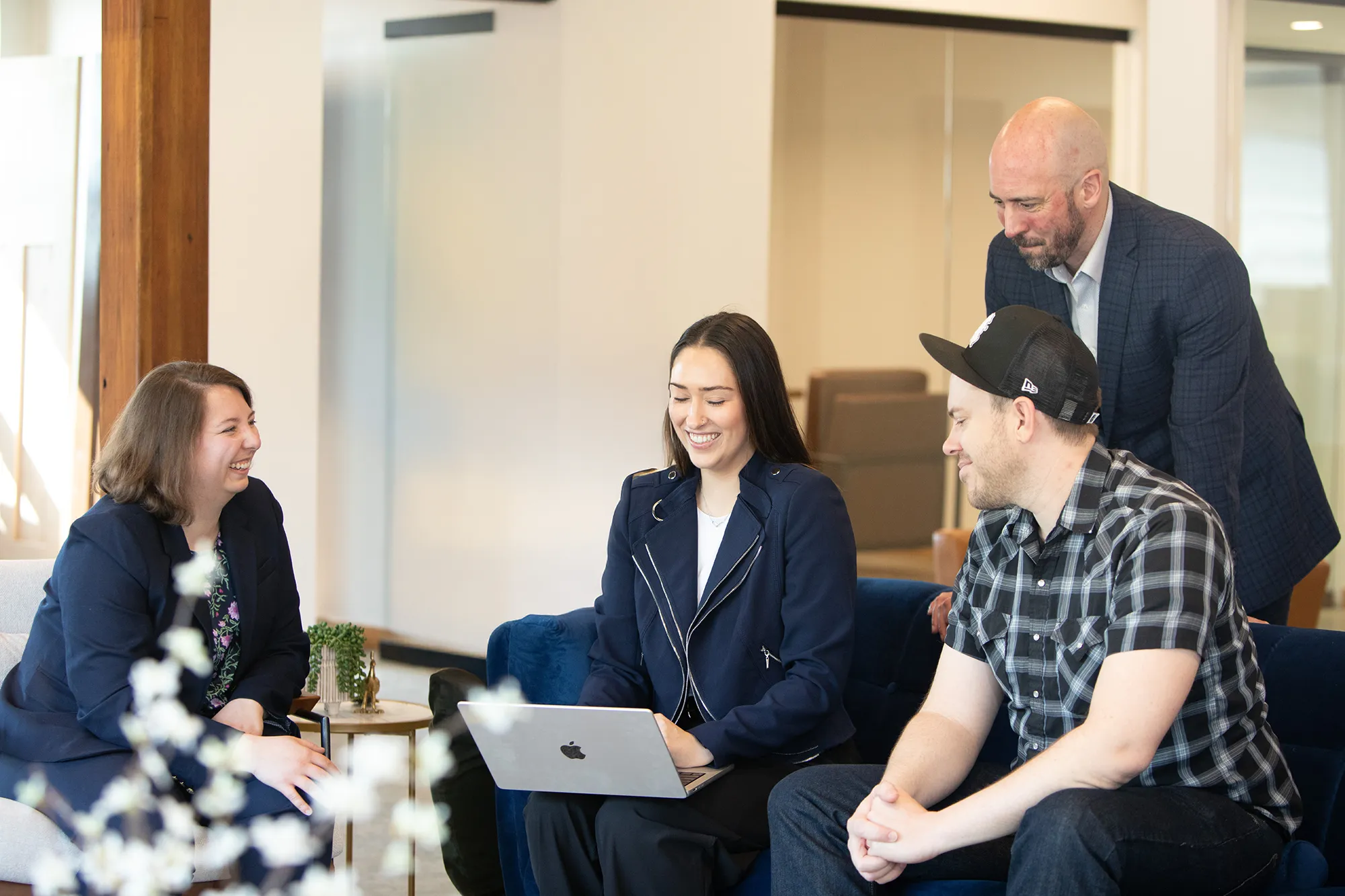 Cleverly Company team members collaborating in a modern office setting. Four people are seated around a laptop, smiling and engaged in discussion, while a fifth person stands behind them. The environment features clean, bright interiors with contemporary furniture, highlighting the agency’s creative and approachable culture.