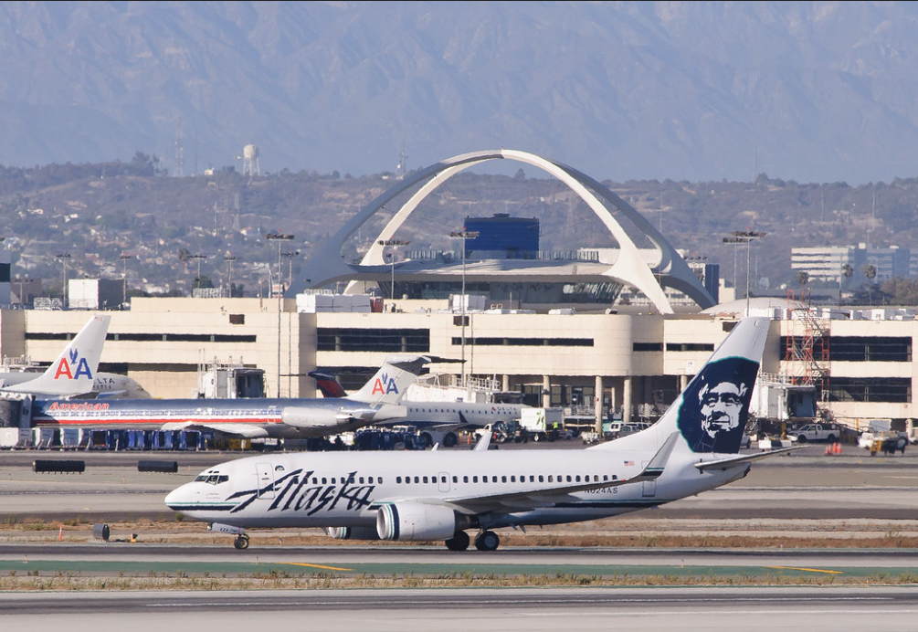Image shows an Alaska Airlines plane taking off with other planes and mountains in the background.