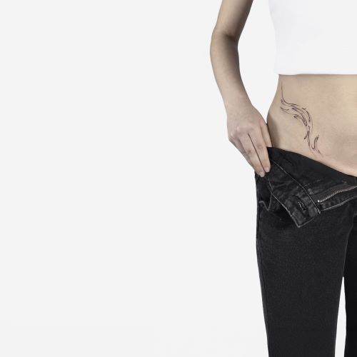 Person iført sort bukser, der trækkes ned for at vise en linjetatovering i abstrakt blækmarmor på venstre hofte og nedre mave.