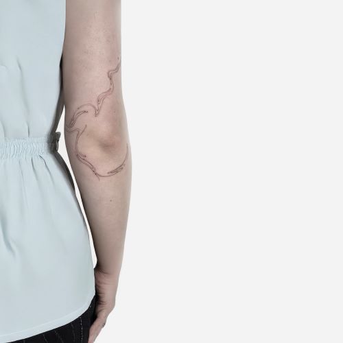 Person iført lyseblå top med elastik i taljen, med en finlinjet, abstrakt tatovering på ydersiden af albuen.