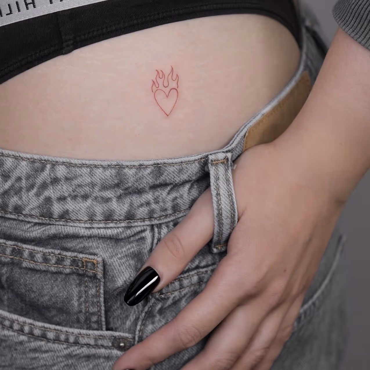 Rød finlinje-tatovering af et hjerte med flammer placeret på den nederste ryg, med en hånd der hviler på taljen iført grå jeans.