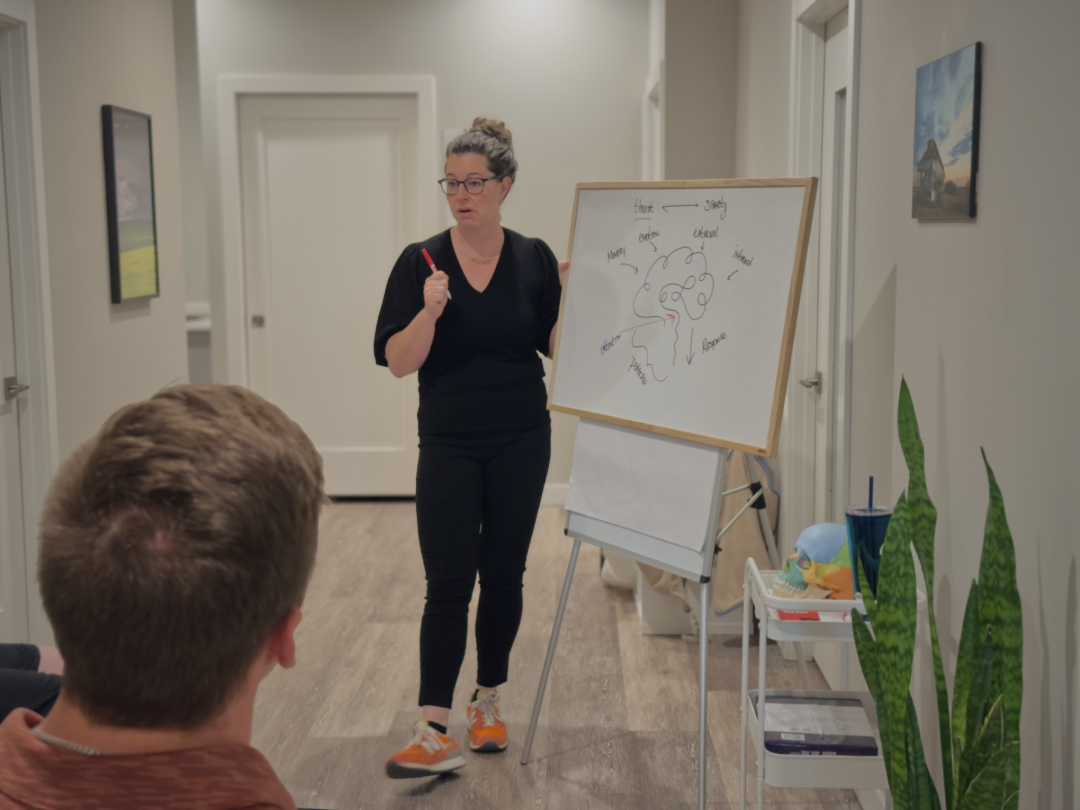 A woman in black scrubs stands next to a whiteboard depicting a stylized drawing of a brain. The woman addresses a class as she speaks.