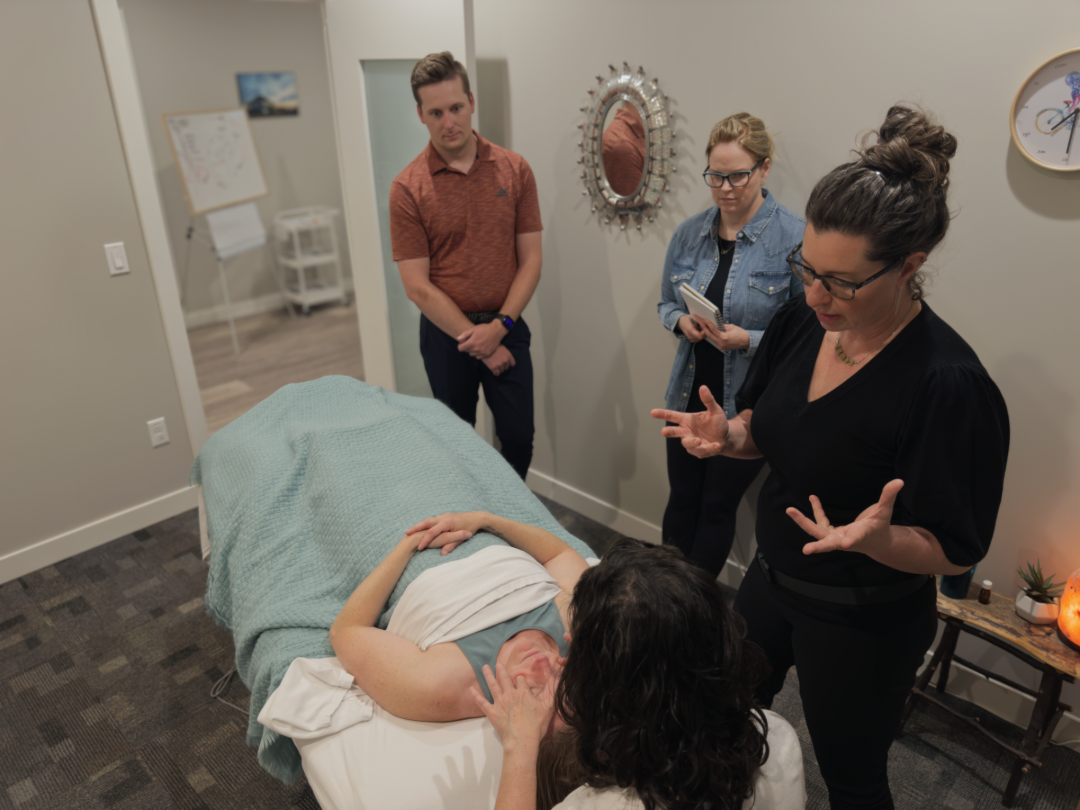 A group of students and a massage therapy instructor stand over a woman laying face up on a massage table. One of the students has both hands on the woman's temple.