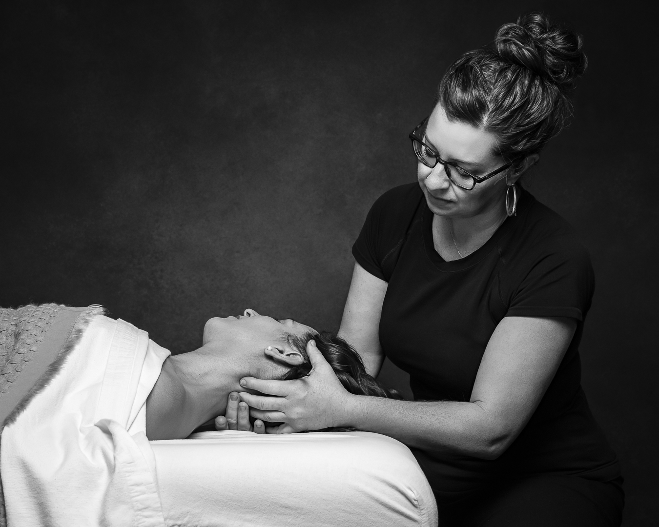 A black an white photo of Alanna Thompson, RMT, with her hands gently on the neck of a young woman on a massage table. 