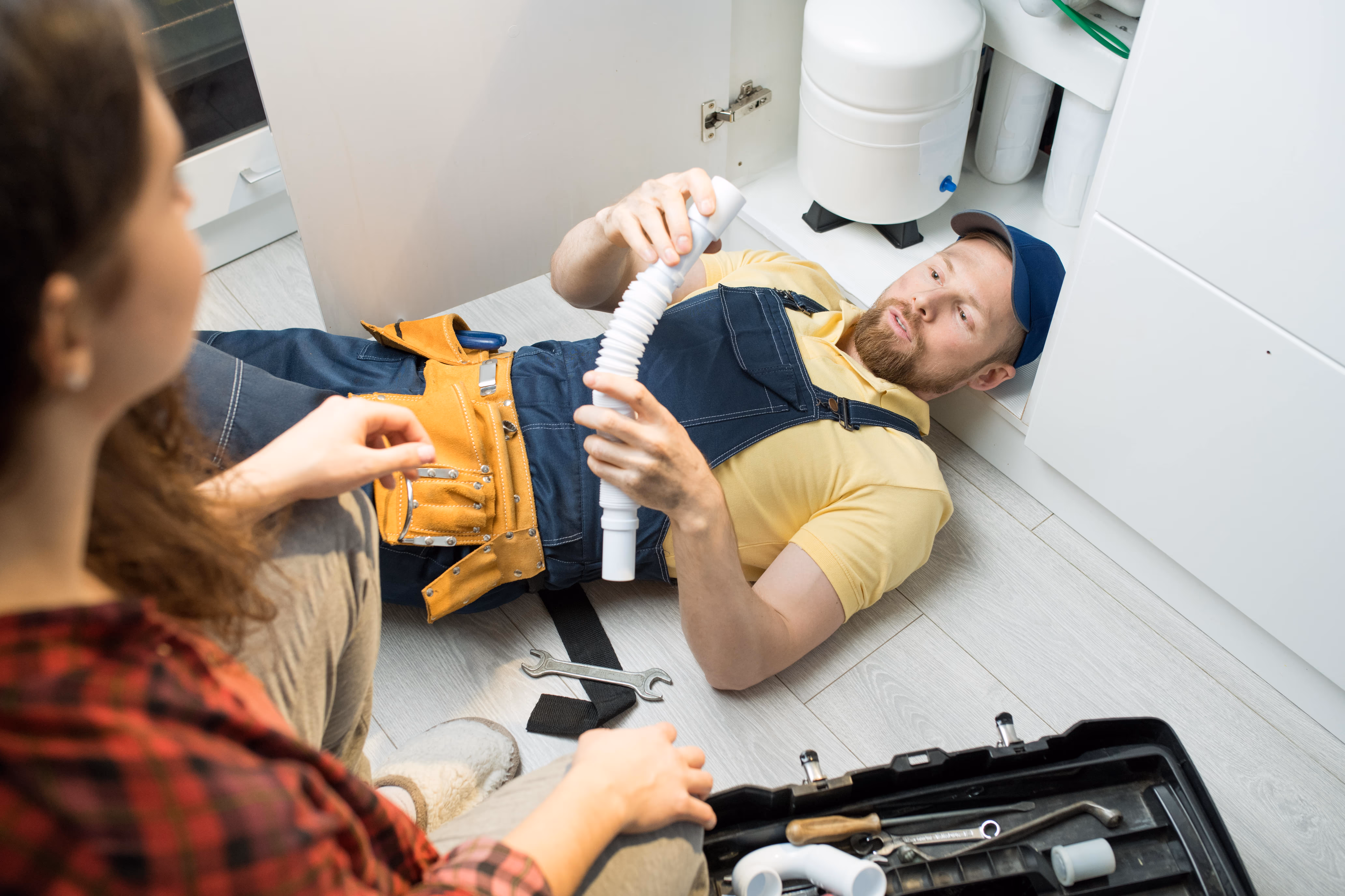 Plumber lying on the floor under a kitchen sink holding a flexible drain pipe while speaking with a female homeowner, surrounded by tools and plumbing parts.