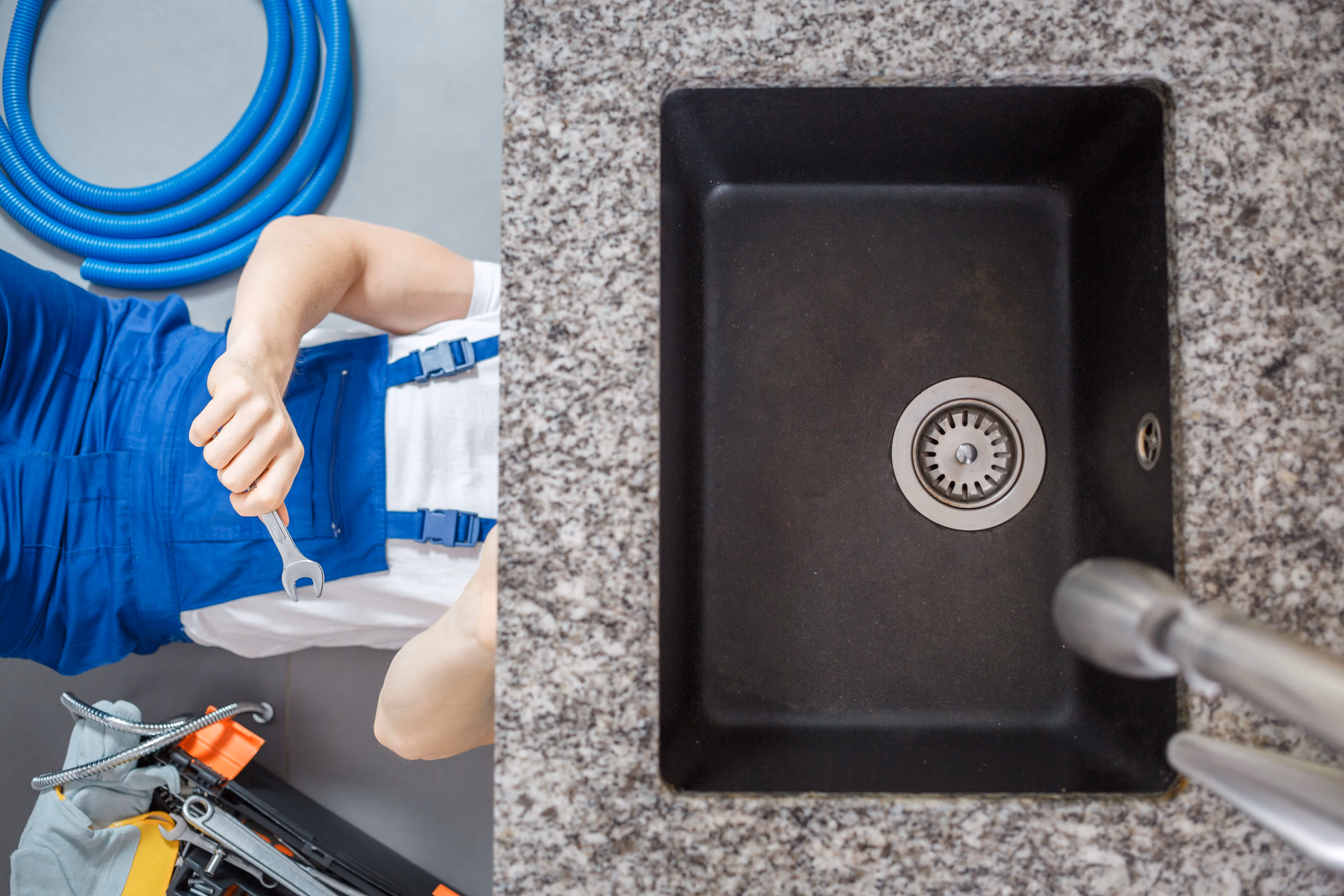 Split view of a plumber lying beneath a kitchen countertop holding a wrench, and a top-down view of a clean black sink with a granite surface.